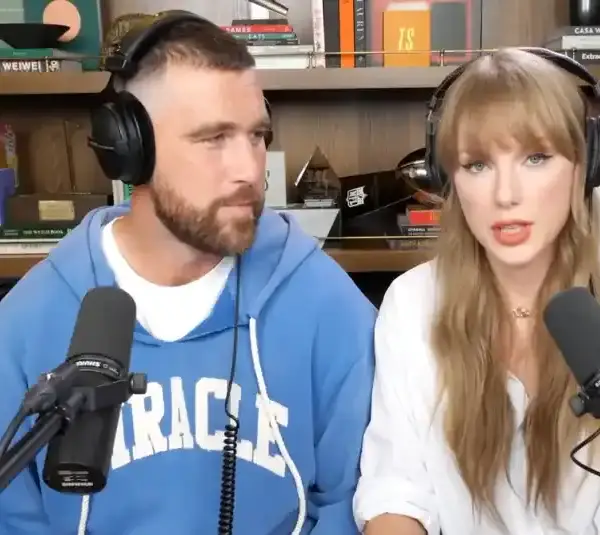 Travis Kelce in a blue hoodie and Taylor Swift in a white shirt, both wearing headphones, speak into microphones on Image of Taylor Swift and Travis Kelce on the New Heights podcast via NHTV Youtube with a bookshelf with books and memorabilia is behind them.