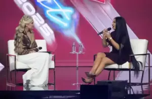 Erika Kirk and Nicki Minaj sit on a stage, each holding a microphone at Turning Point USA's AmericaFest. One wears white pants and a bronze blouse, the other a black dress. A red backdrop reads "2025."