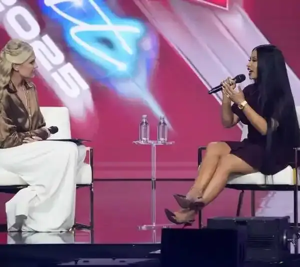 Erika Kirk and Nicki Minaj sit on a stage, each holding a microphone at Turning Point USA's AmericaFest. One wears white pants and a bronze blouse, the other a black dress. A red backdrop reads "2025."