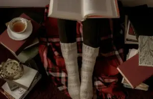 Person reading while sitting on red and black plaid blanket, wearing warm socks, surrounded by books, and a cup of tea., holiday romance