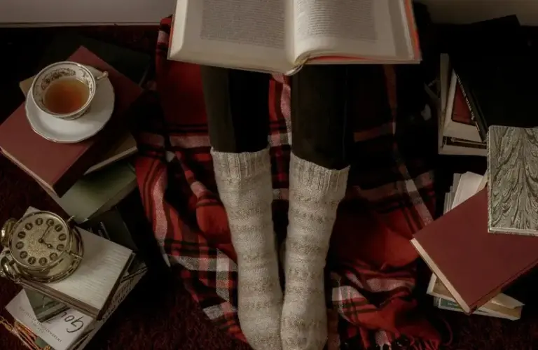 Person reading while sitting on red and black plaid blanket, wearing warm socks, surrounded by books, and a cup of tea., holiday romance