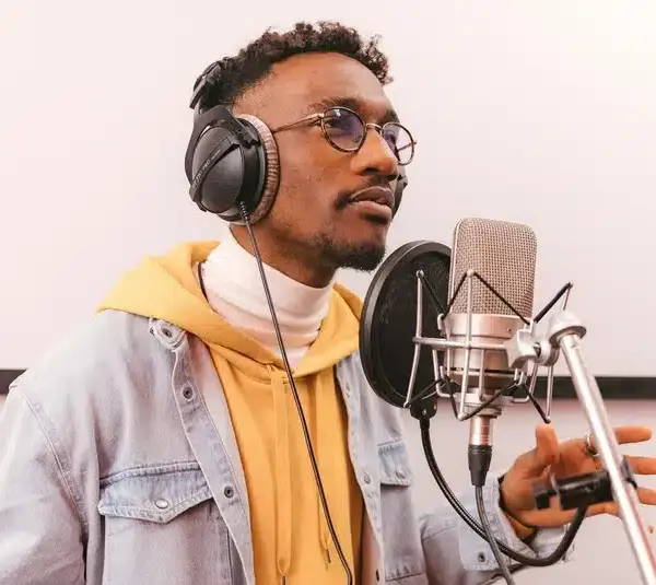 A young man in glasses sings R&B passionately into a microphone. He wears headphones, a yellow hoodie, and a denim jacket, exuding focus and creativity.