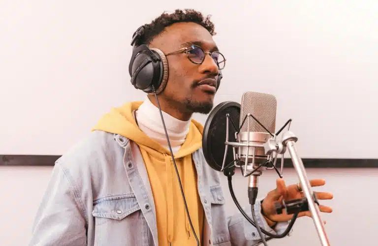 A young man in glasses sings R&B passionately into a microphone. He wears headphones, a yellow hoodie, and a denim jacket, exuding focus and creativity.
