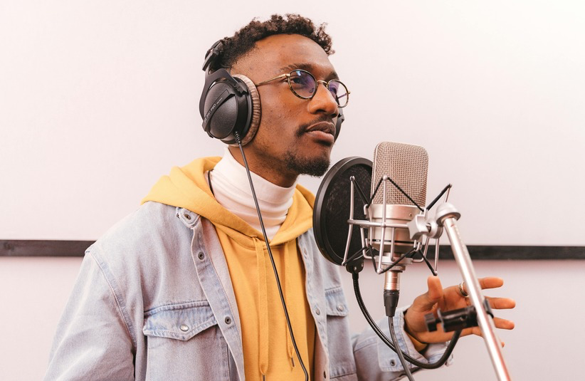 A young man in glasses sings R&B passionately into a microphone. He wears headphones, a yellow hoodie, and a denim jacket, exuding focus and creativity.