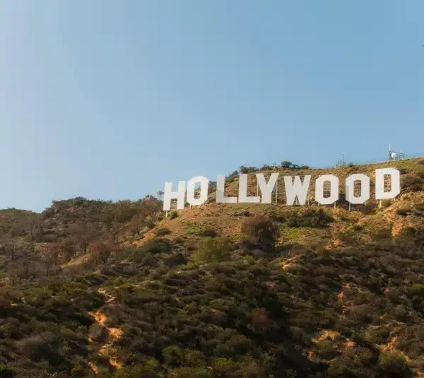 The iconic Hollywood sign is set against a clear blue sky atop a sunlit, grassy hill. It conveys a sense of fame and the film industry's allure.