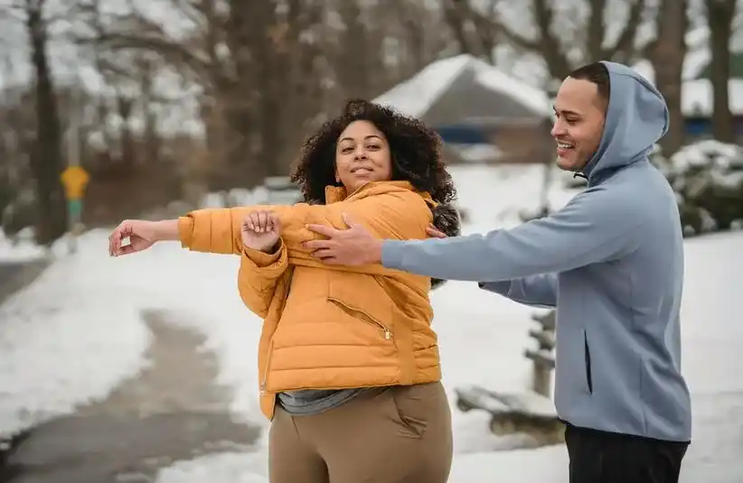 A woman in a yellow jacket playfully stretches her arms while a man in a hooded jacket assists her. They are enjoying a snowy outdoor setting.