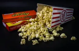 New movie releases in theaters with a tipped popcorn box spills onto a dark surface, surrounded by two colorful candy boxes.
