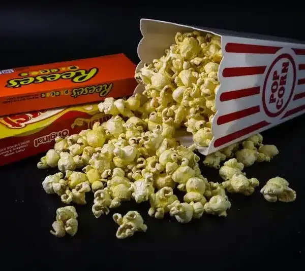 New movie releases in theaters with a tipped popcorn box spills onto a dark surface, surrounded by two colorful candy boxes.