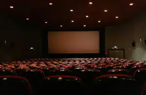 Empty movie theater with rows of red seats facing a large blank screen. Soft ceiling lights create a calm, expectant atmosphere.