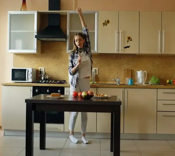 Cheerful young funny woman dancing and singing with ladle while while cooking in the kitchen at home in the morning, holiday