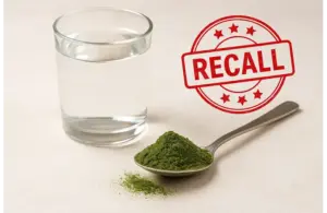 A glass of water and a spoonful of Live It Up Super Greens green powder sitting on a table. A red "RECALL" stamp overlays the scene, suggesting a warning or alert.