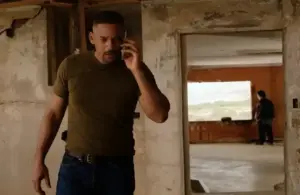 Will Smith a green shirt talks on the phone hearing about his lawsuit, appearing focused, in a dilapidated room with peeling walls. Another person stands near a window in the background.