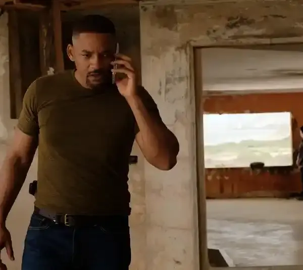 Will Smith a green shirt talks on the phone hearing about his lawsuit, appearing focused, in a dilapidated room with peeling walls. Another person stands near a window in the background.