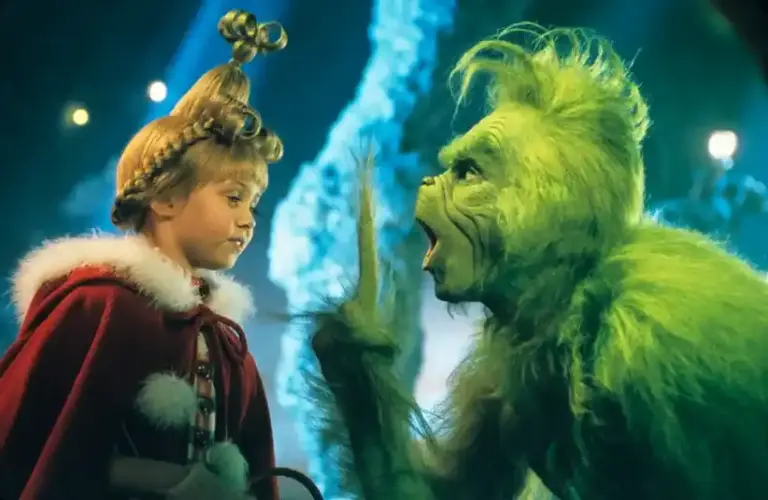 Taylor Momsen, plays young girl with an elaborate hairstyle and red coat faces a green, Jim Carrey, a furry creature gesturing animatedly. The scene is colorful and whimsical, getting ready to The Pretty Reckless rendition of her song in "How the Grinch Stole Christmas," 2000.