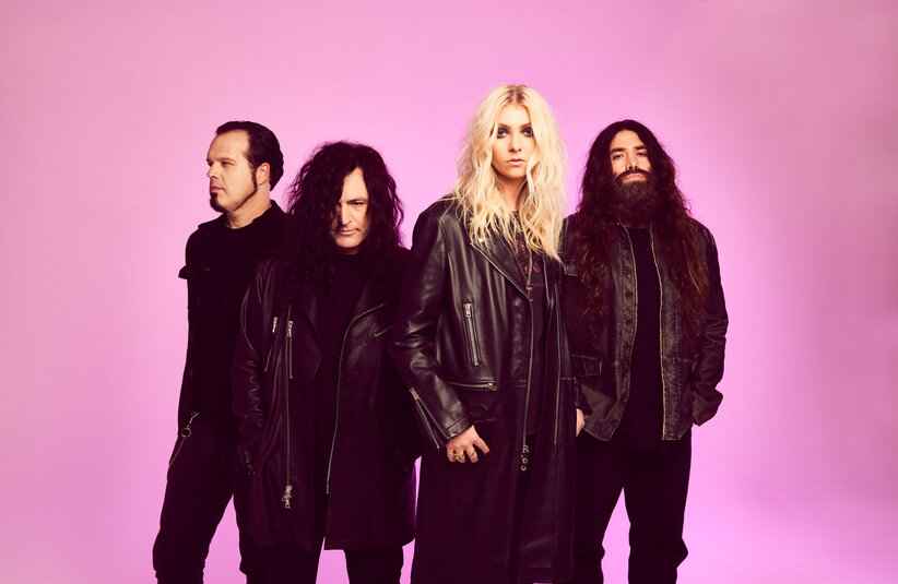 The Pretty Reckless stand against a pink background. They wear black leather jackets and have serious expressions, conveying a confident, rock band vibe.