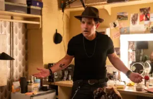 Paul Rudd in a black t-shirt and hat stands in a cluttered dressing room, looking puzzled and unstoppable, holding a red lollipop. Bright lights, casual atmosphere.