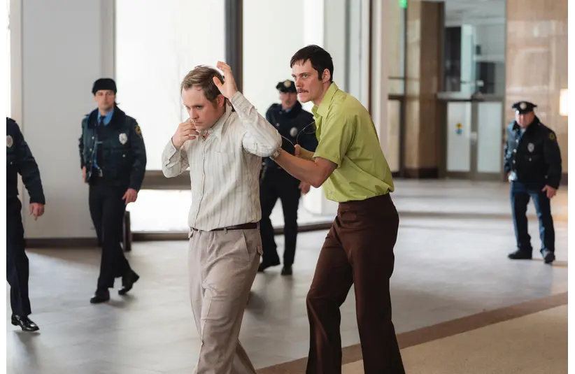 Bill Skarsgård and Dacre Montgomery in Dead Man's Wire (2025), show a man in a green shirt urgently leads another man in a striped shirt with hands raised through a hallway. Police officers watch alertly in the background.