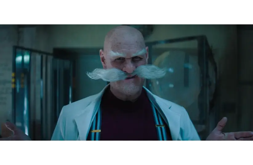 A bald man with a large gray mustache stands in a dimly lit room, wearing a white coat and dark red shirt, conveying a serious and intense mood. Jim Carrey as Dr. Gerald Robotnix, Courtesy of Paramount Pictures and Sega of America, Inc.