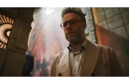 Golden Globes winner, Seth Rogen, in The Studio (2025), in a beige suit, stands in a dimly lit room, exhibiting a serious expression. A colorful, abstract wall mural behind him.
