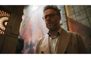 Golden Globes winner, Seth Rogen, in The Studio (2025), in a beige suit, stands in a dimly lit room, exhibiting a serious expression. A colorful, abstract wall mural behind him.