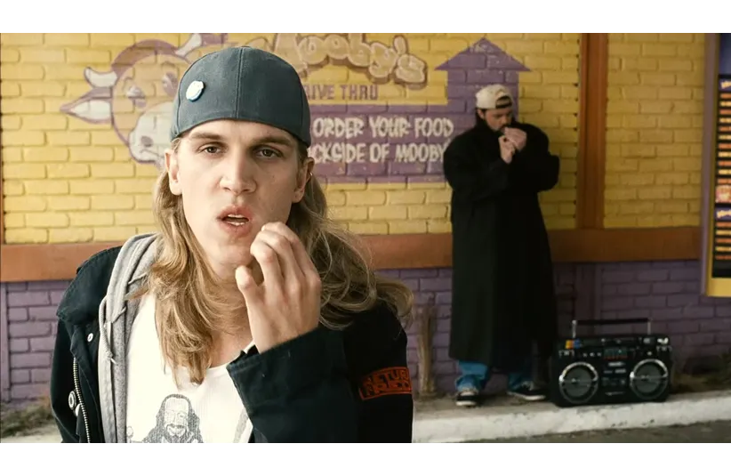 Jay, a young man with long hair, a cap, and casual attire, stands in front of a yellow brick wall with graffiti, looking thoughtful. Another man, Silent Bob, wearing a long coat and a cap, leans against the wall near a boombox.