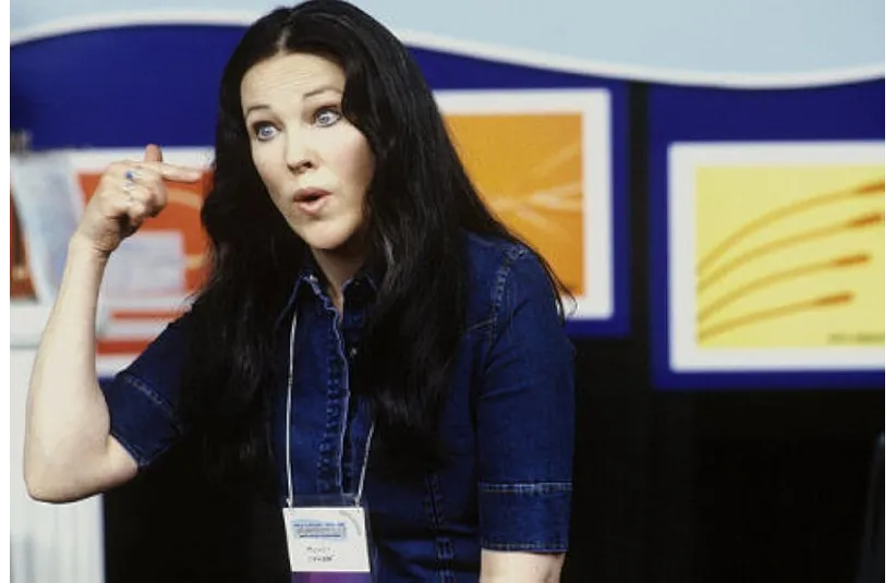 Catherine O'Hara, a woman with long black hair gestures to her head, wearing a denim shirt and name badge. Vibrant abstract art in background. Engaged expression.