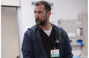 Golden Globes winner, Noah Wyle in The Pitt (2025), wearing a stethoscope and ID badge labeled "DOCTOR," stands in a hospital setting. He has a thoughtful expression, conveying professionalism.