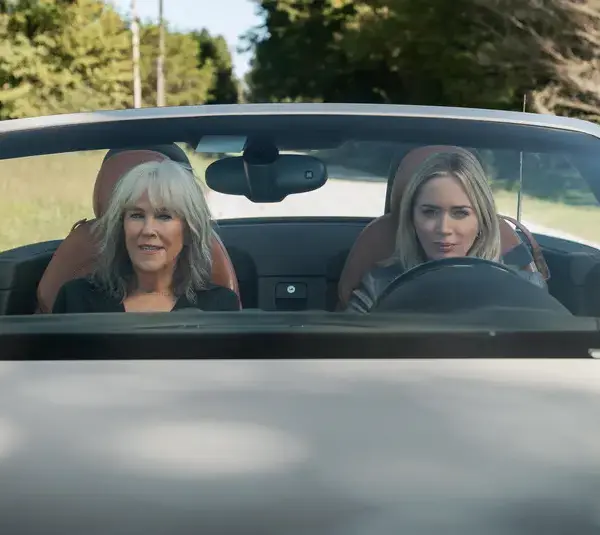 Catherine O'Hara and Emily Blunt sit in a convertible car, driving down a country road surrounded by greenery. They appear relaxed and content, enjoying the sunny day.