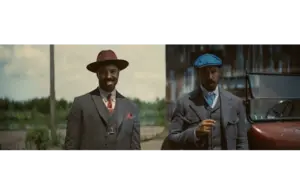 Michael B. Jordan in Golden Globes-winning "Sinners (2025)" as Smoke & Stack in vintage suits stand outdoors near a classic car. The man on the left wears a burgundy hat, while the right wears a blue cap. The scene feels nostalgic.