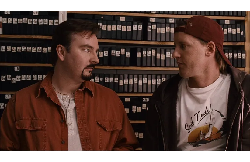 Two men, Dante and Randal, stand facing each other in a room lined with shelves full of small boxes. The man on the left wears an orange shirt and looks inquisitive, while the man on the right, in a cap and "Sail Nude!" shirt, appears casual. The tone is conversational and engaged.