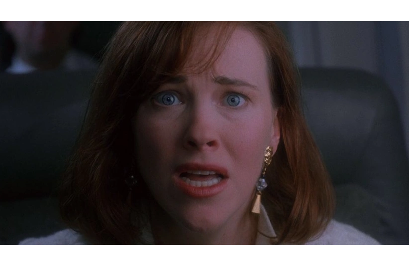Catherine O'Hara, a woman with red hair looks shocked and worried, her mouth open and eyes wide. She wears gold earrings and sits in a dimly lit setting, conveying tension. She is about to say "KEVIN!" in Home Alone.