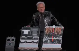 NVIDIA CEO Jensen Huang speaking live at CES 2026, he is in a shiny black jacket presents two large computer hardware components on stage. The components are detailed, with visible wires and circuits.