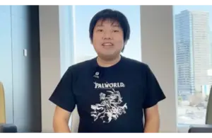 Poketpair CEO, Takuro Microbe, in a Palworld t-shirt, smiles in a bright office with urban buildings visible through large windows. The atmosphere is casual and friendly.