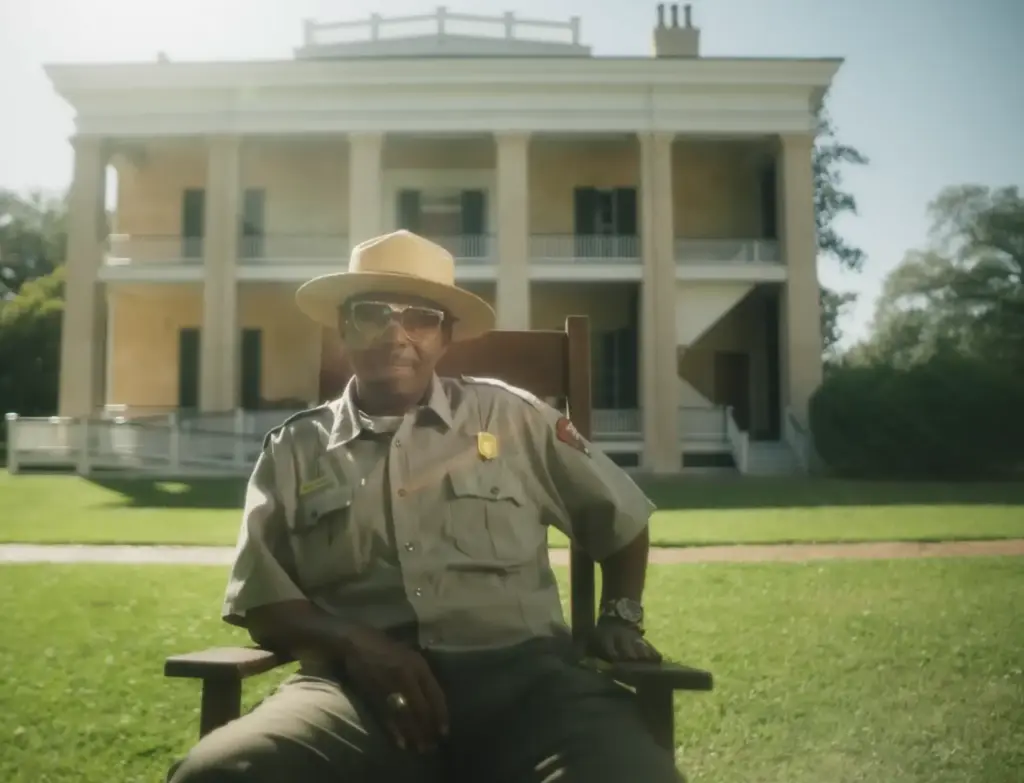 A park ranger in uniform sits relaxed on a chair in front of a historic mansion. The sunlit scene conveys a sense of pride and tranquility in the film "Natchez."
