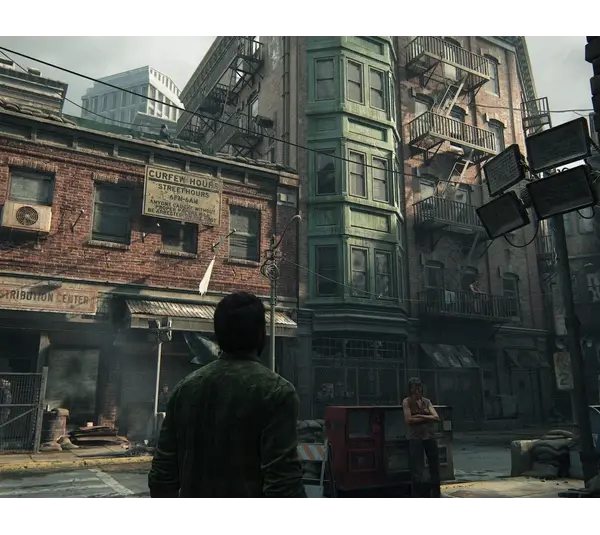 Joel, in a green jacket, stands in an abandoned, run-down city street. Surrounding are old brick buildings with fire escapes, creating a tense, post-apocalyptic atmosphere in The Last of Us