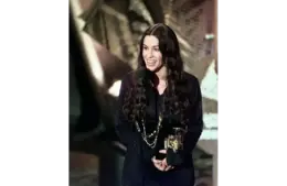 Alanis Morissette is a 2026 inductee for the Songwriters Hall of Fame. She has long dark hair, smiles joyfully while holding a trophy in one hand and speaking at a microphone, set against an abstract background.