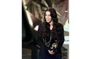 Alanis Morissette is a 2026 inductee for the Songwriters Hall of Fame. She has long dark hair, smiles joyfully while holding a trophy in one hand and speaking at a microphone, set against an abstract background.