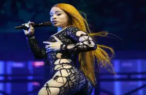 Ice Spice with long orange hair sings passionately on stage, wearing a black lace outfit with intricate cut-outs, conveying energy and confidence.