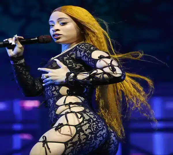 Ice Spice with long orange hair sings passionately on stage, wearing a black lace outfit with intricate cut-outs, conveying energy and confidence.