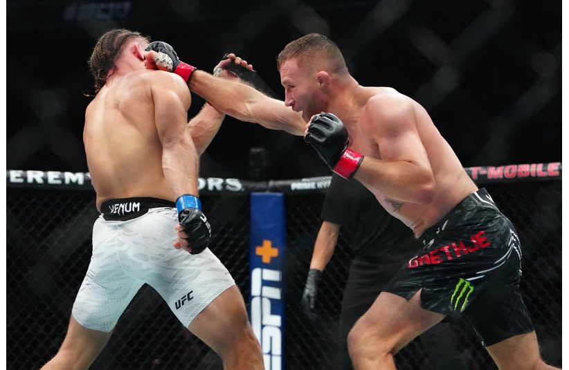 Gaethje, one of the two mixed martial artists are in a UFC fight. One fighter lands a punch while the other blocks. The referee watches, and the crowd is blurred behind the cage.