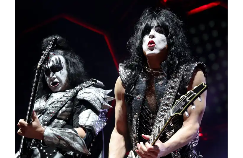 Gene Simmons and Paul Stanley from KISS. The two performers in extravagant costumes play electric guitars on stage. Their faces are painted white with black designs, creating a dramatic, theatrical look.
