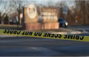 Crime scene tape creates a barrier in front of a blurred outdoor scene featuring a parked car and a sign. The atmosphere feels tense and serious. Recently, a fake ice agent robbed a home.