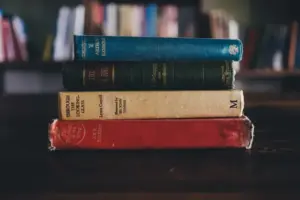 A stack of four vintage books with worn covers sits on a dark wooden table, set against a blurred background of a bookshelf, evoking a nostalgic library feel. Could they be part of the International Booker Prize list of 2026?
