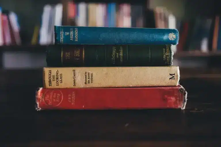 A stack of four vintage books with worn covers sits on a dark wooden table, set against a blurred background of a bookshelf, evoking a nostalgic library feel. Could they be part of the International Booker Prize list of 2026?