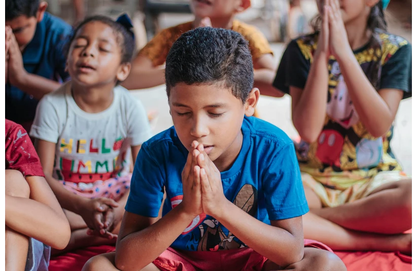 A group of kids, children, eyes closed, sit cross-legged, palms clasped in prayer. The atmosphere is calm and focused, suggesting a meditative setting.