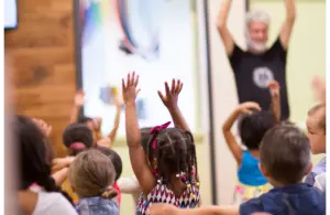 A group of young children with diverse hairstyles eagerly raise their arms in a lively children's church classroom. An adult leads them, creating a joyful, energetic atmosphere.