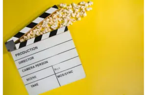 New movie releases in cinemas and theaters this weekend are shown by a film clapperboard with popcorn spilling out, lying on a vibrant yellow background. The open clapper gives a playful feel, symbolizing cinema and entertainment.