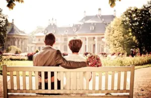 Image of a couple in wedding attire sits on Valentine's Day embracing and facing a grand resort. The scene is bathed in warm sunlight, evoking romance and serenity.