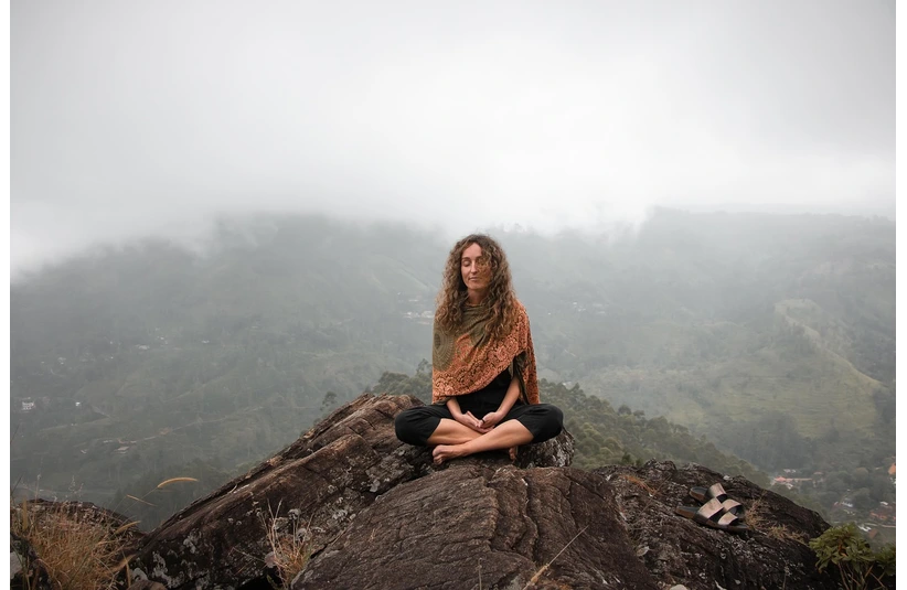 Person meditating on a rocky cliff, wearing a shawl, surrounded by misty mountains. Serene and peaceful atmosphere with sandals nearby.