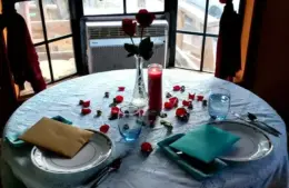image of Romantic table setting by a window, featuring a round table with a white lace cloth, red rose petals, a candle, two plates, glasses, and a vase with roses for Valentine dinners